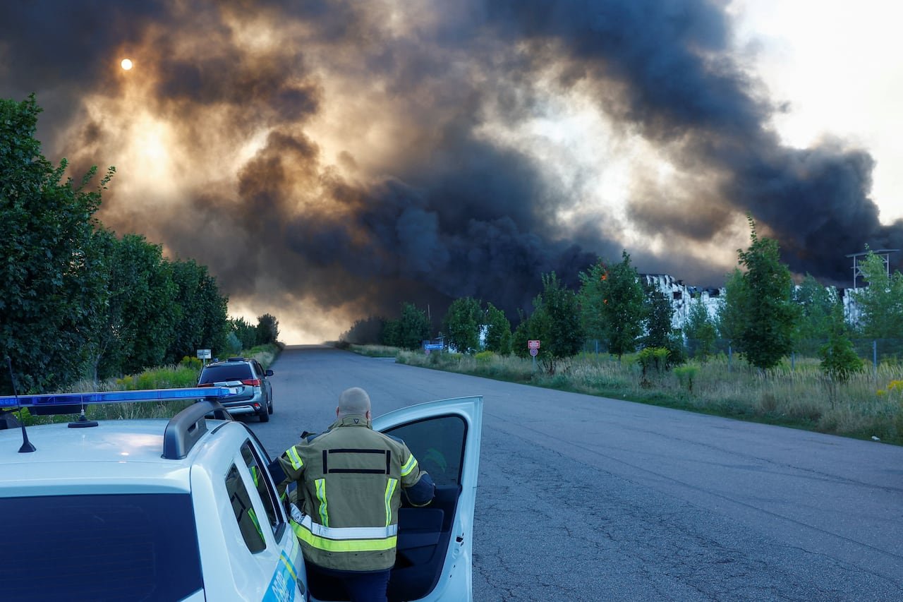 default-466 A firefighter with his back to the camera gets out of the passenger side of a vehicle as off in the distance of a rural area, large black clouds of smoke fill the sky near a series of low-rise structures.